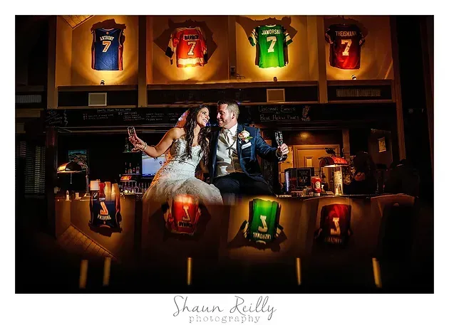 Couple on a bar counter surrounded by glowing framed sports jerseys, warm nightclub lighting.