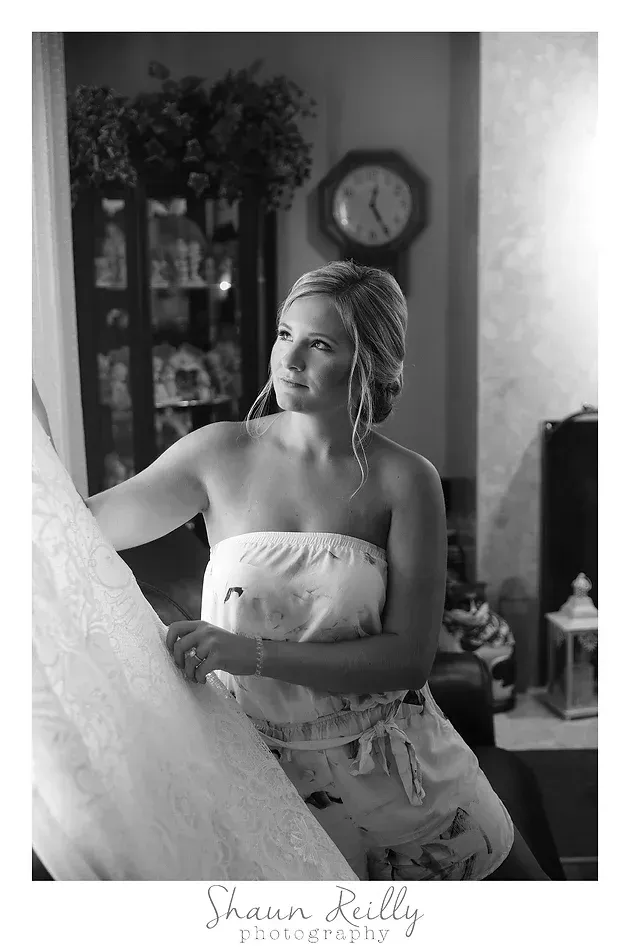 Woman in a strapless dress standing by a window, looking out in a softly lit room.
