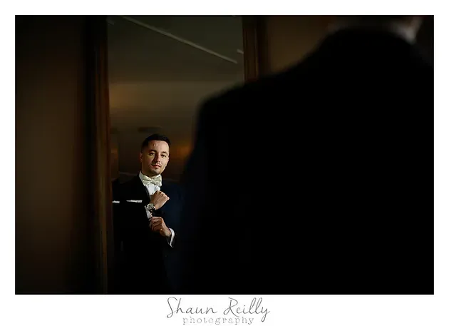 Man in a mirror adjusting his suit, with a blurred dark foreground and warm indoor lighting