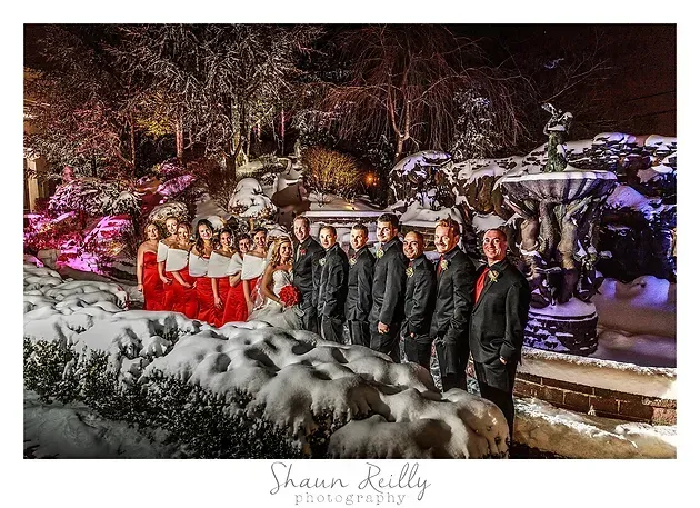 Formal group portrait outdoors at night, with red-dressed women and men in dark suits in a snowy, lit garden