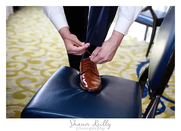 Person lacing a brown dress shoe while sitting on a blue chair in a patterned room