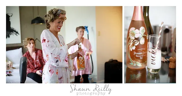 Woman in floral robe holding drink at a bridal prep table with bridesmaid and decorated bottle
