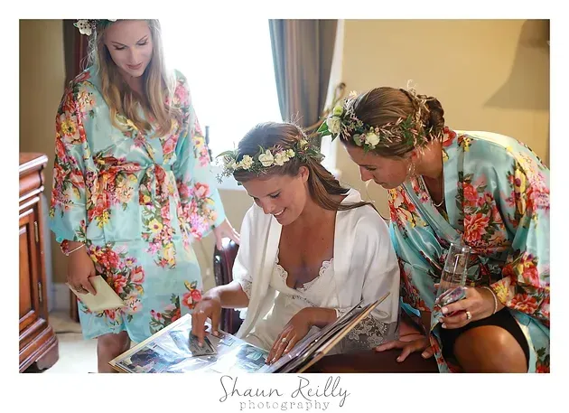 Three women in floral robes and flower crowns prepare together indoors by a window