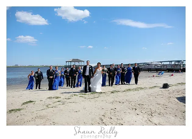 Wedding party posing on a beach under a blue sky, with bride and groom centered among guests in blue attire