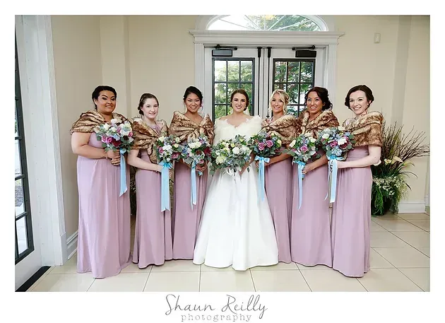 Bride in white gown with five bridesmaids in lavender dresses, holding bouquets indoors