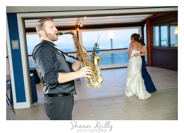 Saxophonist in dark shirt plays indoors while a bride in a white gown stands in the background.