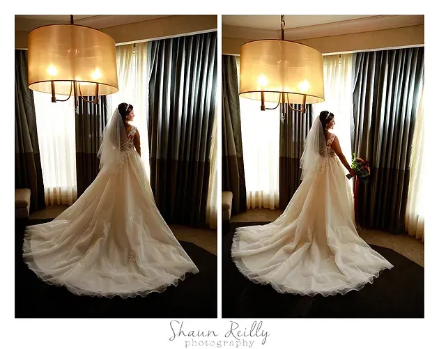 Bride in a white gown standing by a window, holding a bouquet, with a long train in a softly lit room