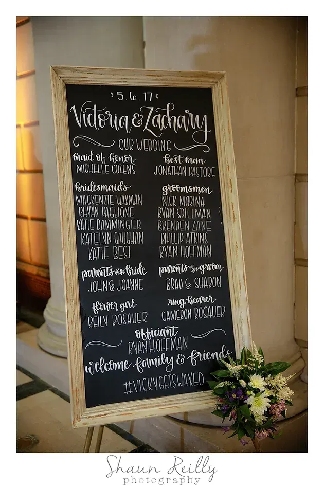 Black wedding welcome sign on an easel with white calligraphy and a small flower arrangement beside it