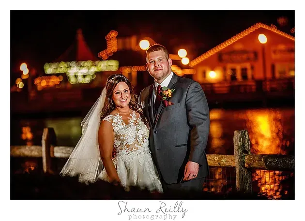 Bride and groom posing at night by a lit waterfront venue, with warm lights reflecting on the water