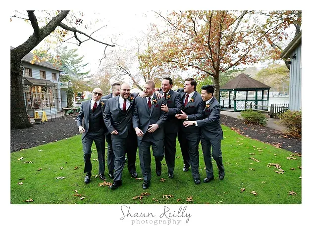 Group of men in suits posing outdoors on a lawn with autumn trees and a gazebo behind them