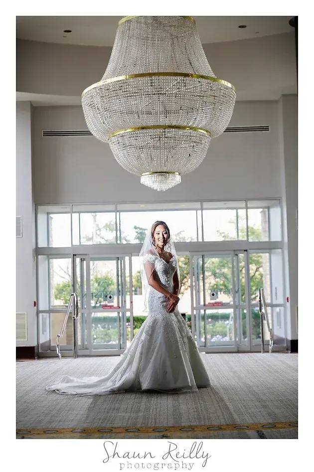 Bride in a white gown holding a bouquet beneath a large chandelier in a bright lobby