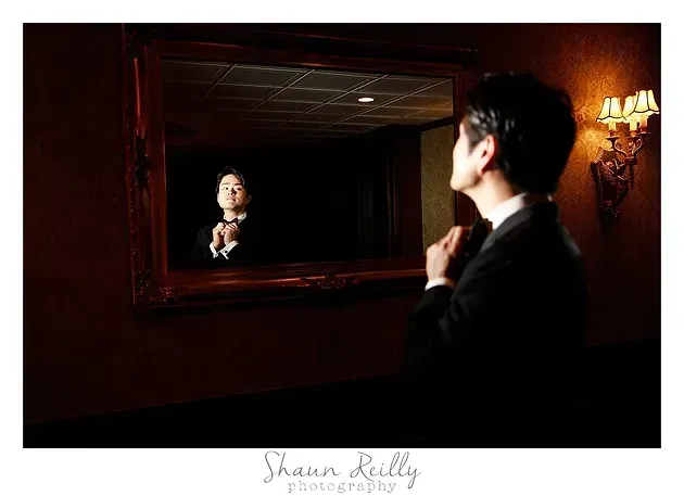 Person in a dark suit adjusts their tie while looking into a mirror in a dimly lit room.