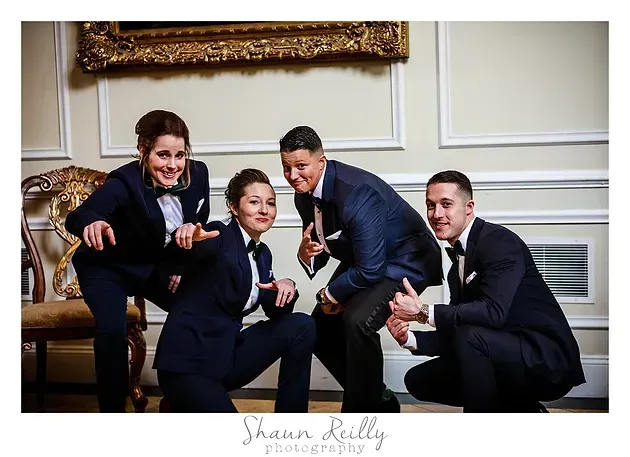 Four men in navy suits pose smiling indoors, seated on a couch in a bright room.