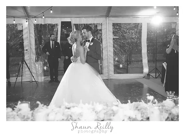Bride and groom slow dancing at their wedding reception under string lights, surrounded by guests and decor