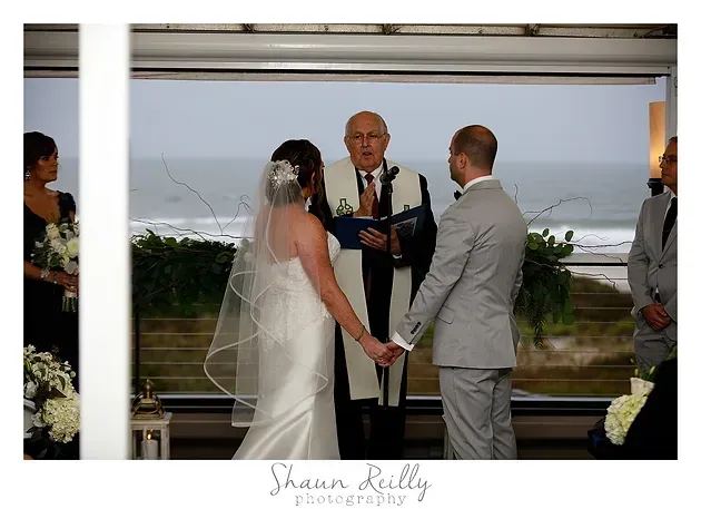 Wedding ceremony by the ocean, bride in white and groom in gray holding hands as officiant speaks