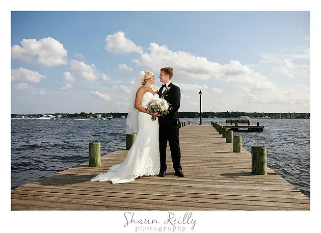 Bride and groom embracing on a wooden dock by the water under a partly cloudy sky