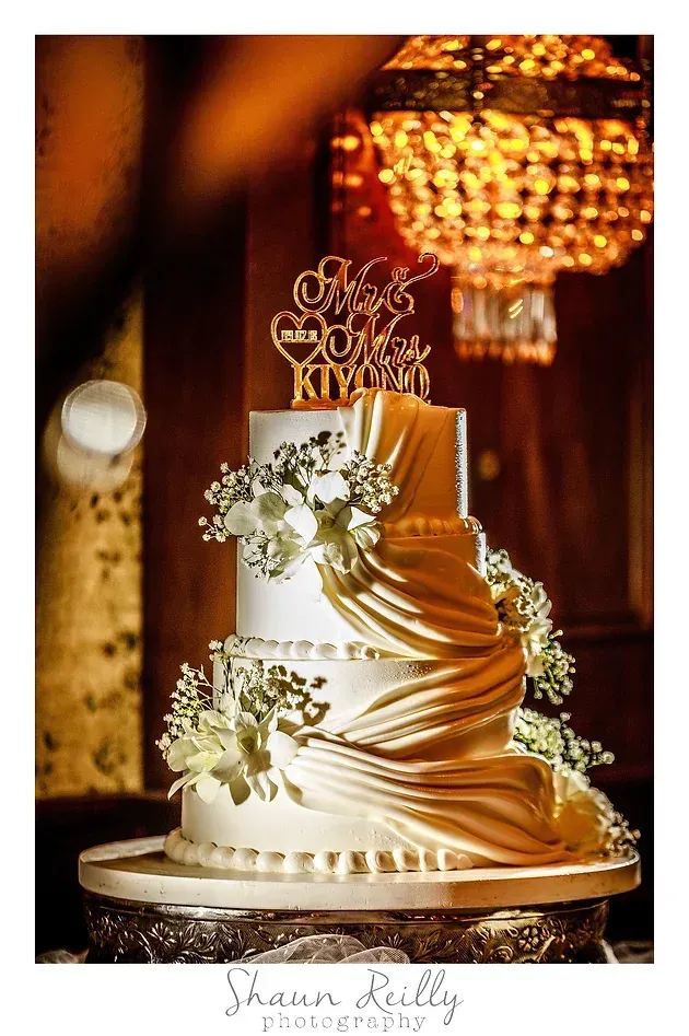 Elegant white-and-gold tiered wedding cake with floral accents and a chandelier backdrop