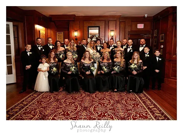 Wedding party posed in a wood-paneled room, women in black and gold dresses, men in tuxedos, flower girl in white.