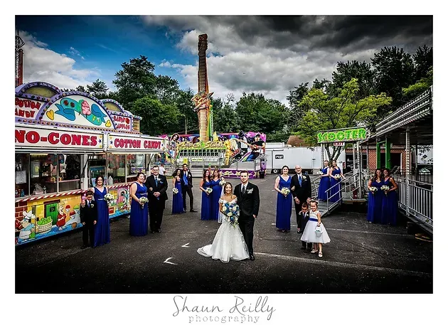 Wedding party in blue attire poses in a colorful outdoor fairground under stormy skies