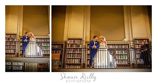 Couple in blue and white wedding attire posing on a library balcony, with bookshelves behind them.