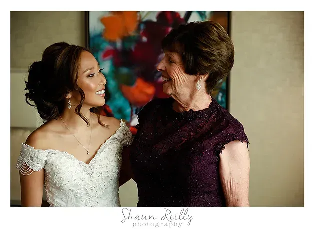 Two women smiling at each other, one in a white lace dress and one in a purple dress, indoors with colorful artwork