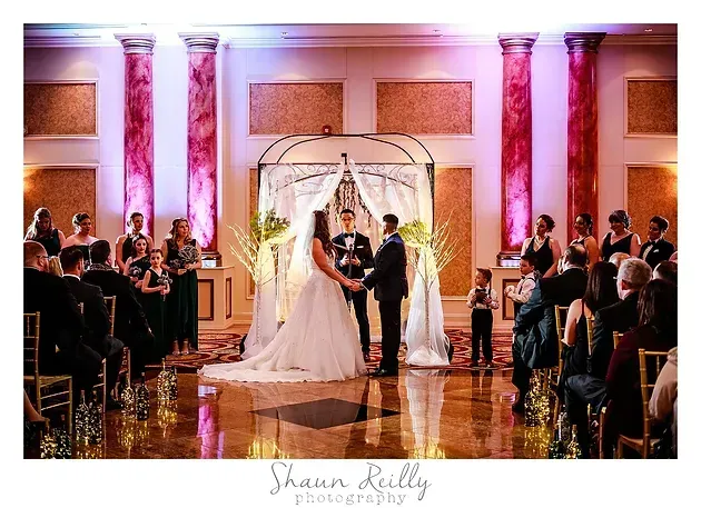Wedding couple under a floral arch in a warmly lit ballroom, guests seated on both sides