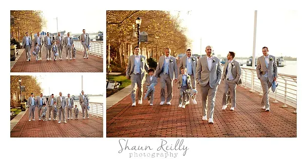 Wedding party walking down a brick path beside autumn trees and water, with one groom in a blue suit in front