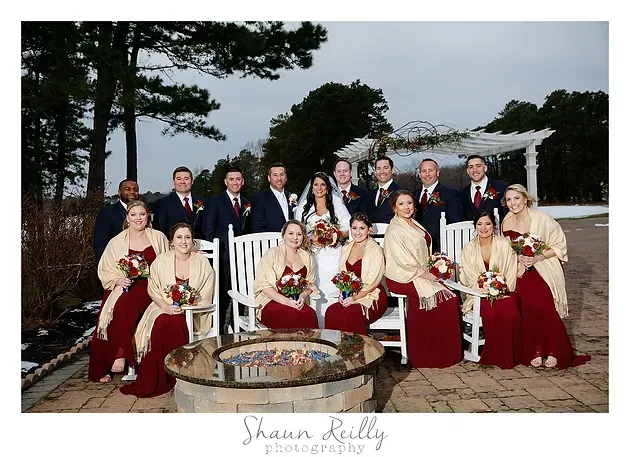 Wedding party posed outdoors by a fountain, with men in dark suits and women in red dresses and cream shawls.