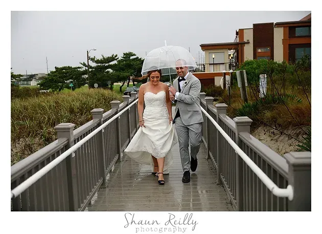 Bride and groom walking hand in hand under a clear umbrella on a rainy bridge
