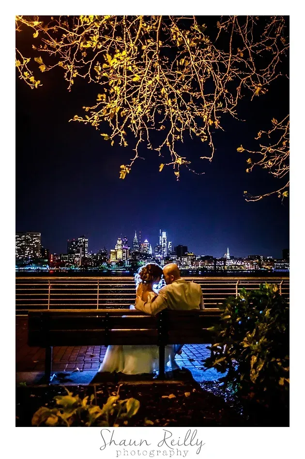Couple embracing on a bench at night with city lights across the water and tree branches overhead