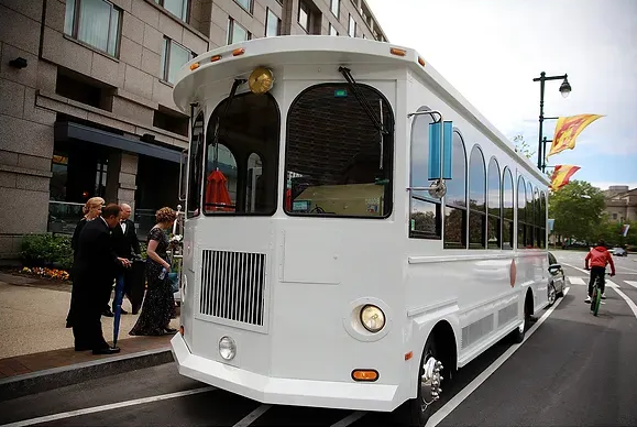 White open-air tour tram on a city street with people boarding near tall buildings and colorful flags.