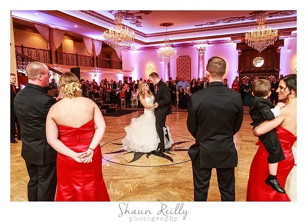 Bride and groom dancing at a wedding reception under pink lighting, guests watching nearby