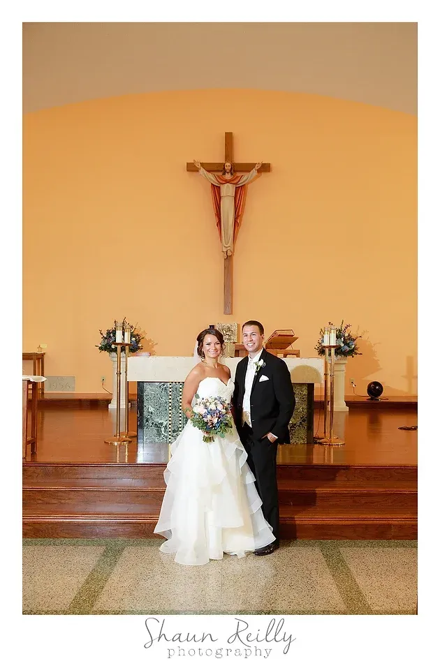 Bride and groom standing at a church altar beneath a crucifix, with white flowers and warm orange walls