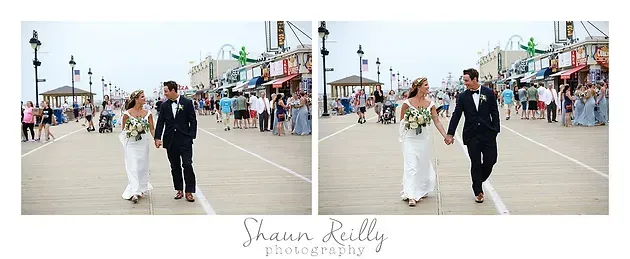 Couple in wedding attire walking hand in hand on a boardwalk, with shops and a beach in the background.