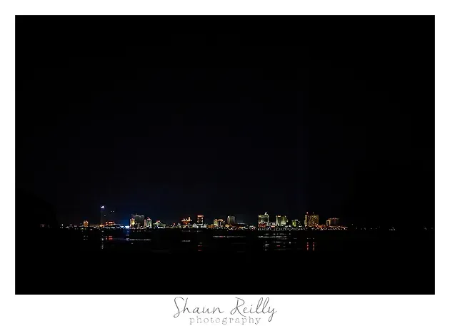 Night city skyline across dark water, with colorful lights glowing along the horizon.