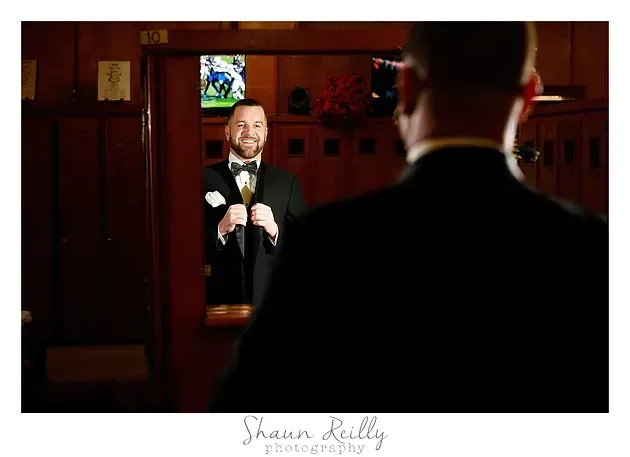 Man in tuxedo speaking at a microphone, seen in a mirror with another figure in the foreground.