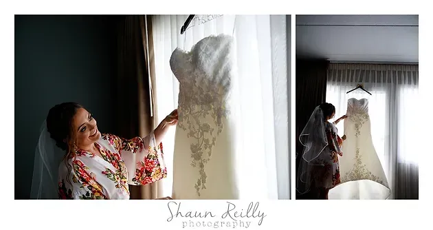 Bride adjusts wedding dress by window, with white gown hanging and a person in reflection beside it