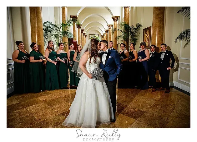 Wedding couple kissing in a grand hallway, bridesmaids and groomsmen lined behind them