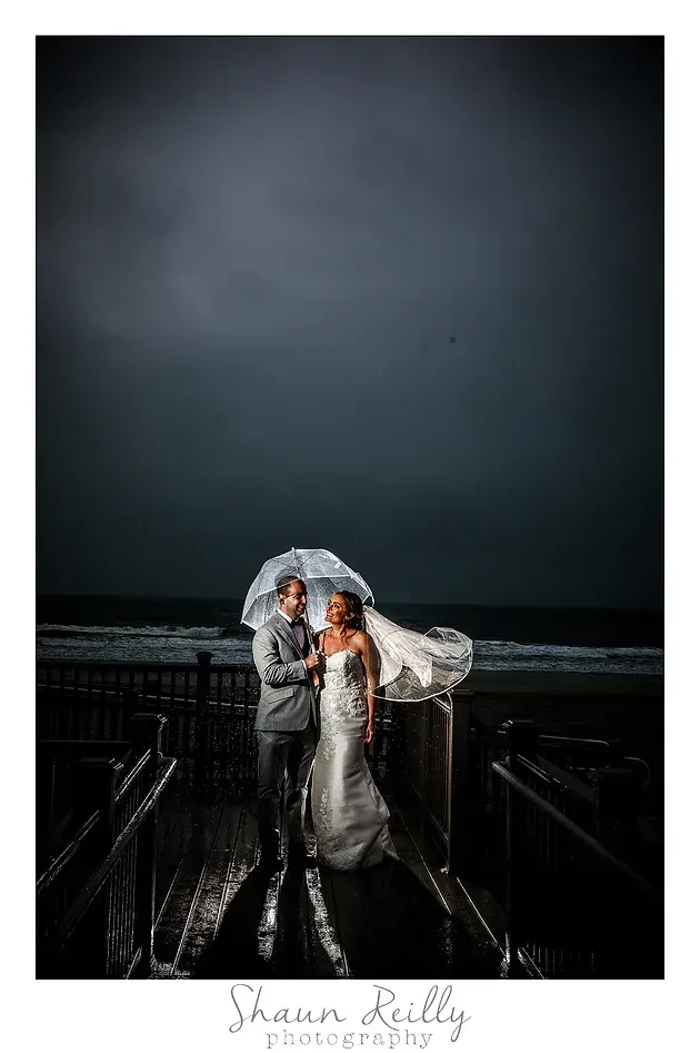 Bride and groom under a clear umbrella on a dark rainy dock, with stormy sky and ocean behind them