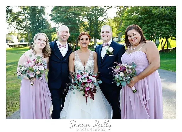 Wedding party posing outdoors in pastel dresses and suits, holding bouquets under trees.