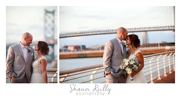 Couple in wedding attire embracing by a waterfront bridge at sunset