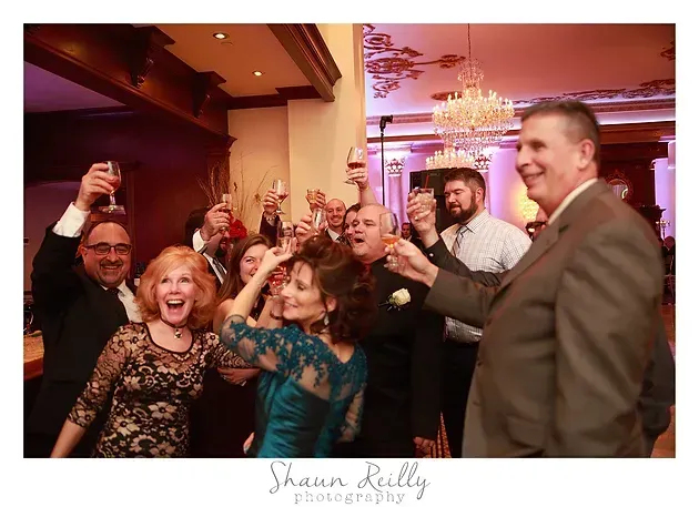 Guests cheer and raise drinks at a lively indoor party with warm lighting and a chandelier