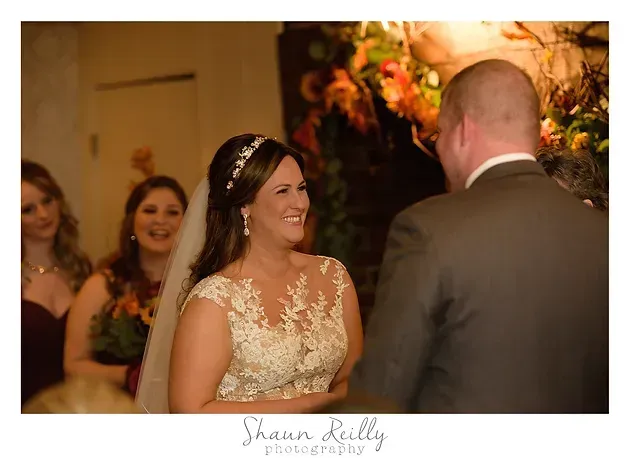 Bride smiling at groom during wedding reception, with guests and warm floral decor in background