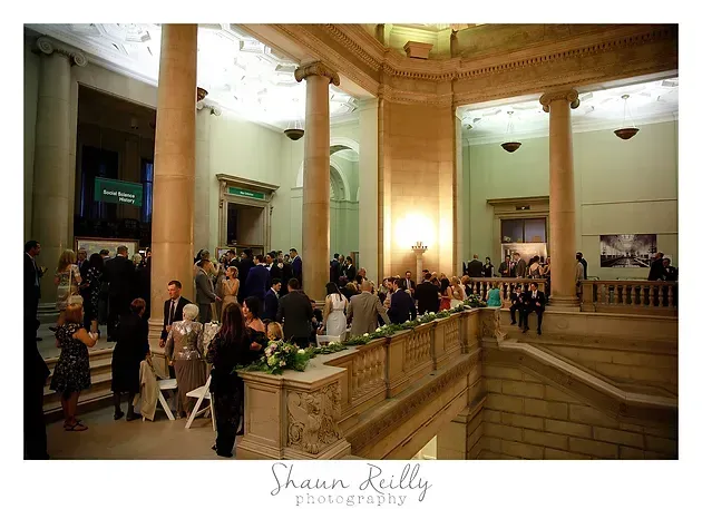 Guests mingling in a grand columned hall with warm lighting and a staircase at the right.
