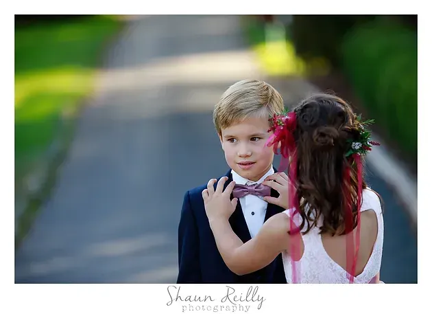 Two children in formal wear facing each other on a blurred outdoor path, with a red flower in one child's hair