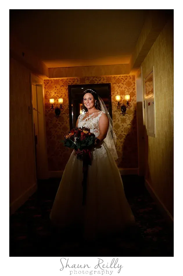 Bride in white gown holding bouquet in a dimly lit hallway, framed by warm wall sconces.