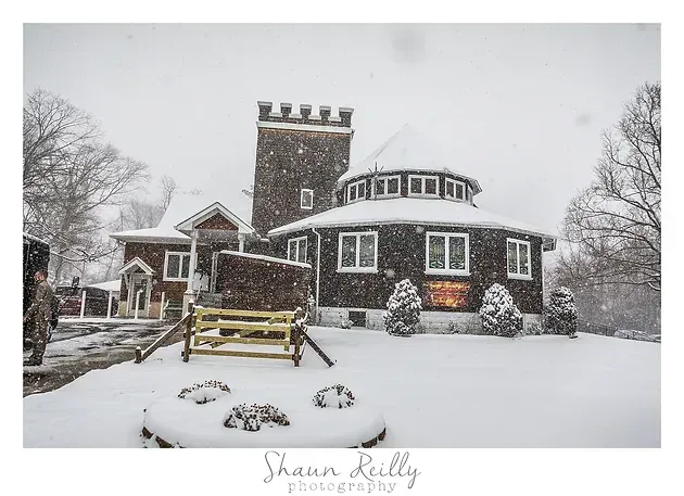 Snow-covered stone house with a tower and curved front, surrounded by trees in a winter storm.