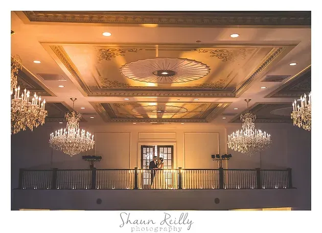 Elegant ballroom ceiling with ornate chandeliers and warm lighting above a balcony
