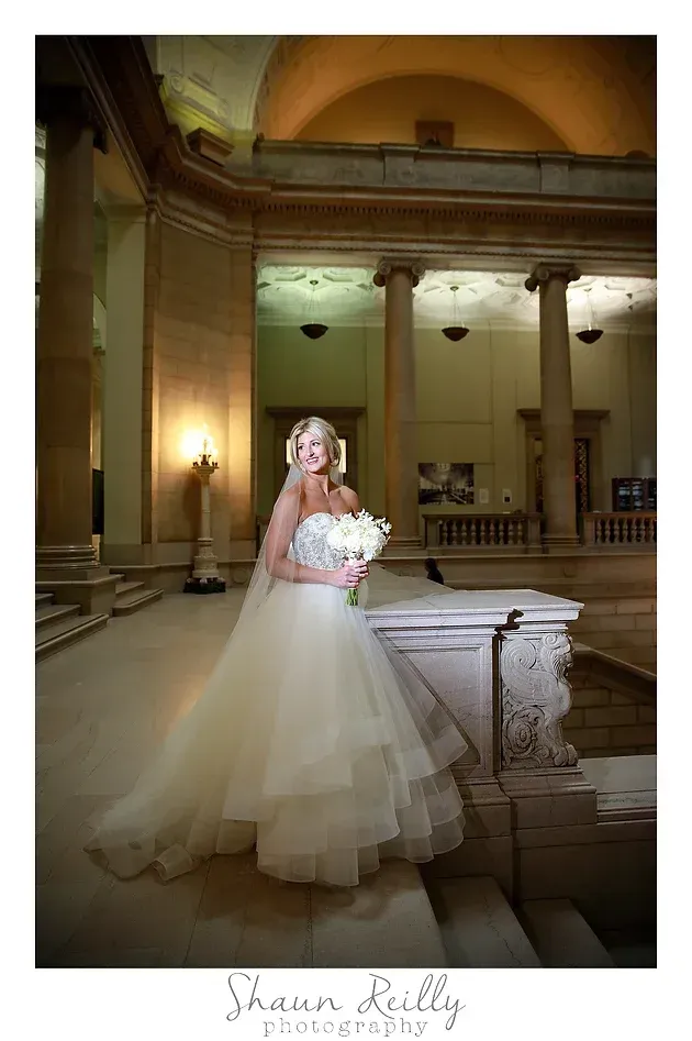Bride in a white gown holding a bouquet in an ornate hall, standing by a marble railing.