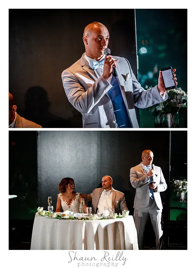 Man in a white blazer speaking into a microphone at a wedding reception while guests sit at the head table